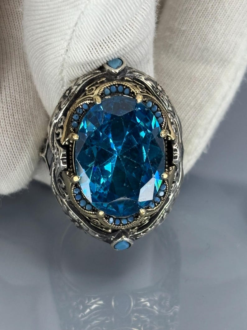 Gloved close-up front view of the oval aquamarine blue stone set in a 925 sterling silver Ottoman-style men’s ring on gray background.