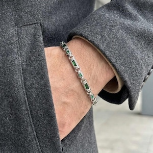 Puede incluir: Una pulsera de plata con piedras preciosas verdes rectangulares y redondas. La pulsera se lleva en la muñeca, con un abrigo gris en el fondo. La pulsera es una joya.