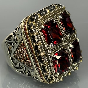 Puede incluir: Un anillo de plata con un engaste cuadrado que presenta cuatro piedras preciosas rojas. El anillo tiene intrincados detalles de filigrana y una banda texturizada.