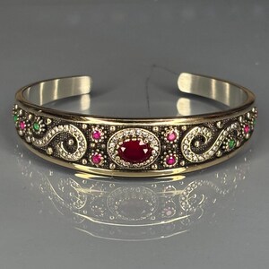 Könnte beinhalten: Ein silber-goldenes Armreif-Armband mit einem ovalen, tiefroten Edelstein in der Mitte, umgeben von weißen Steinen. Das Armband ist mit kleineren rosa und grünen Edelsteinen und aufwendigen Schnörkeln verziert.