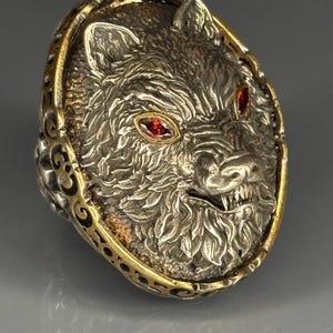 Può includere: Anello argentato con una testa di lupo dettagliata con occhi di gemme rosse. L'anello ha un bordo dorato con un design ornato. La pelliccia del lupo è strutturata.