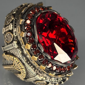 Op de afbeelding: Een sierlijke ring met een grote, ovaal geslepen, rode edelsteen. De ring heeft een zilverkleurige band met goudkleurige details en kleinere rode edelstenen. Het ontwerp is ingewikkeld en gedetailleerd, met een vintage esthetiek.