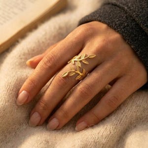 May include: A gold-colored ring with a leaf design. The ring wraps around the finger with two leaf-shaped elements. The ring is worn on a hand with manicured nails, set against a soft, neutral background.