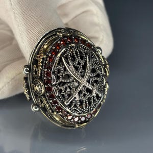 Op de afbeelding: Versierde zilveren ring met een gedetailleerd filigraanontwerp. De ring heeft een centraal ontwerp van gekruiste zwaarden, omringd door kleine rode edelstenen. De ring heeft een vintage, antieke uitstraling.