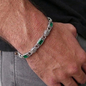 Può includere: Bracciale in argento con pietre verdi rettangolari e maglie ornate in filigrana. Il bracciale è mostrato su un braccio, evidenziando il suo design dettagliato e il contrasto cromatico. Un accessorio alla moda.