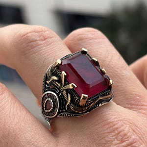 May include: A silver ring featuring a large, rectangular, deep red gemstone. The ring has intricate gold and black detailing around the stone and on the band, with a circular accent of smaller stones.