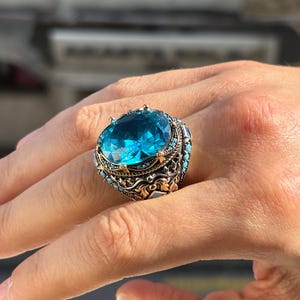 May include: A large, ornate ring featuring a vibrant, oval-cut, light blue gemstone. The ring has a detailed, antique-style setting with smaller turquoise-colored stones and intricate metalwork. The ring is worn on a finger.