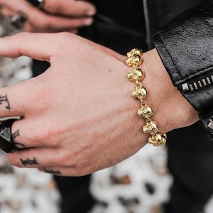 May include: A gold bracelet with a repeating skull design. The bracelet is worn on a wrist, with a black leather jacket visible. The skulls are detailed and the bracelet appears to be made of metal.