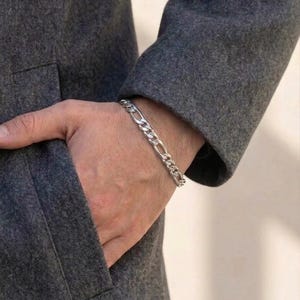 May include: A silver Figaro chain bracelet is displayed on a wrist, set against a grey coat. The bracelet features a repeating pattern of three small links and one elongated link. The coat is a solid, medium grey colour.