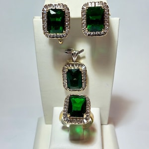 Könnte beinhalten: Ein Schmuckset mit Ring, Anhänger und Ohrringen. Jedes Stück hat einen großen, rechteckigen, smaragdgrünen Stein, der von kleineren, klaren Steinen umgeben ist. Ring und Ohrringe haben eine goldfarbene Fassung. Der Schmuck wird auf einem weißen Ständer präsentiert.