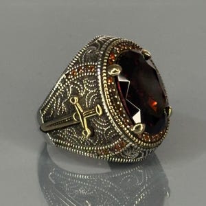 Könnte beinhalten: Ein silberner Ring mit einem großen, dunkelbraunen Edelstein, der von kleineren, orangefarbenen Edelsteinen umgeben ist. Der Ring hat ein filigranes Design mit einem Schwert und einem Kreuz.