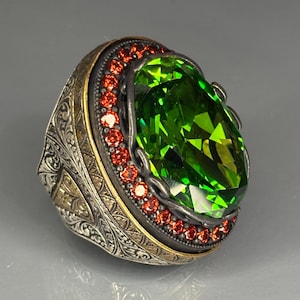 Könnte beinhalten: Ein aufwendiger Ring mit einem großen, leuchtend grünen Edelstein. Der Ring hat ein silbernes Band mit detaillierten Gravuren und ein goldfarbenes Innenband. Kleine rote Edelsteine umgeben den großen grünen Stein.