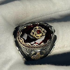 以下が含まれることがあります： 金色の透かし細工のデザインに、大きな赤い宝石がセットされた銀の指輪。指輪は黒色の背景に金色の文字が刻まれており、精巧な作りになっている。