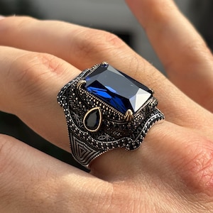 Puede incluir: Anillo plateado con una gran piedra preciosa rectangular de color azul oscuro como pieza central. El anillo está adornado con piedras preciosas negras más pequeñas y detalles intrincados alrededor de la banda y el engaste. Una piedra azul en forma de lágrima más pequeña está engastada en el lateral.