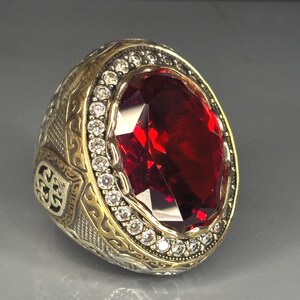 Può includere: Un anello ornato con una grande gemma rosso scuro tagliata a forma ovale. L'anello ha una fascia color oro con intricati motivi incisi e un bordo di piccole gemme chiare che circondano la pietra centrale. L'anello è posto su uno sfondo grigio.