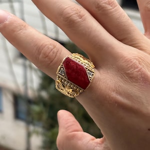 Könnte beinhalten: Ein goldfarbener Ring mit einem großen, roten, rechteckigen Stein in der Mitte. Der Ring hat ein verziertes Filigran-Design mit kleinen, klaren Akzentsteinen auf beiden Seiten des zentralen Steins. Der Ring befindet sich an einem Finger.