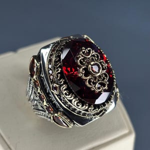 May include: A silver ring with a large, faceted, oval-shaped, deep red gemstone as the centerpiece. The ring features intricate filigree and smaller red gemstones around the main stone. The design includes gold accents, creating a luxurious appearance.