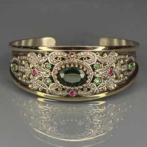 Op de afbeelding: Goudkleurige manchetarmband met een ovale, donkergroene edelsteen in het midden. De armband heeft ingewikkelde filigraan en is versierd met kleine rode en groene edelstenen en heldere kristallen. Het ontwerp is symmetrisch en sierlijk.