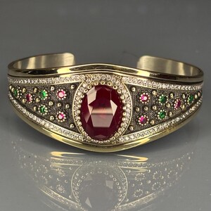 Puede incluir: Brazalete de puño dorado con una gran gema de rubí ovalada en el centro, rodeada de pequeños diamantes blancos. La banda está adornada con pequeños acentos de rubí, esmeralda y diamantes.