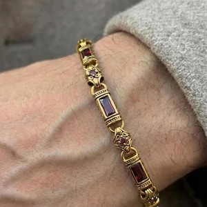 May include: A gold-colored bracelet with rectangular and round red gemstones. The bracelet is made of linked segments with ornate detailing. The bracelet is worn on a wrist, with a gray sleeve in the background.