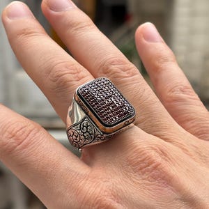 Könnte beinhalten: Ein silberner Ring mit rechteckiger Oberfläche, besetzt mit kleinen, dunkelroten Steinen. Der Ring hat ein verziertes, graviertes Design an den Seiten und einen goldfarbenen Rand um die Steinfassung. Der Ring wird am Finger getragen.