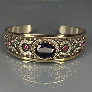 Puede incluir: Una pulsera de puño de plata y oro con una piedra preciosa ovalada de color azul oscuro en el centro. La pulsera presenta detalles intrincados con pequeñas piedras preciosas rojas, verdes y transparentes. El diseño es simétrico y adornado.