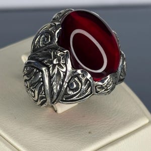 Könnte beinhalten: Ein silberner Ring mit einem verzierten Design und einem großen, ovalen, roten Edelstein.