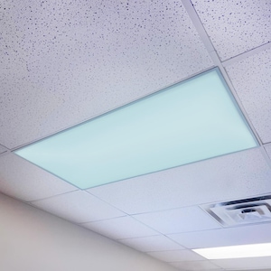 May include: A rectangular, recessed fluorescent light fixture installed in a drop ceiling. The light panel emits a soft, cool white glow, framed by a thin, silver-colored border. The ceiling tiles are white with a speckled texture.