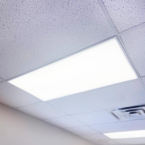 May include: A bright, rectangular LED ceiling light fixture, recessed into a white, textured ceiling. The light is surrounded by a thin, silver frame. The ceiling tiles have a speckled pattern. The image is taken from a low angle.