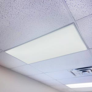 May include: A rectangular, bright white LED panel light installed in a drop ceiling. The light is surrounded by a silver frame and is flush with the white ceiling tiles. The ceiling tiles have a speckled texture.