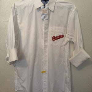May include: A crisp white long-sleeve button-down shirt featuring a pointed collar and a chest pocket. The pocket displays the red embroidered text 'Sooners'. A branded tag hangs from the collar, and the sleeves are rolled up to the elbows.