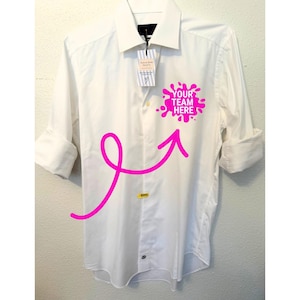 May include: A white long-sleeve button-down shirt featuring a bright pink ink-blot graphic on the chest with the text "YOUR TEAM HERE" in white. A large pink arrow graphic points toward the ink-blot, and a product tag hangs from the collar.