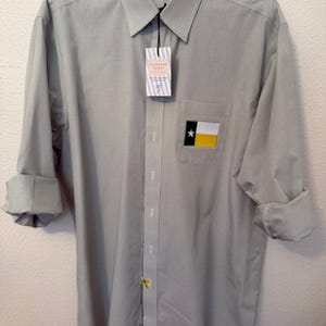 May include: A light gray long-sleeve button-down shirt with a fine grid pattern. The shirt features a chest pocket with a rectangular patch displaying a black, white, and yellow flag with a white star. A white tag with text hangs from the collar.