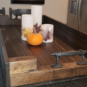 May include: A wooden serving tray with a dark brown stain and a natural wood finish. The tray has two metal handles and is decorated with fall-themed candles and a small pumpkin.