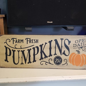 Puede incluir: Un letrero de madera con el texto "Farm Fresh Pumpkins 25c Open Daily" en letras negras sobre un fondo beige. El letrero presenta una calabaza estilizada con un tallo naranja y un cuerpo naranja.