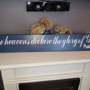 May include: A long, rectangular navy blue wooden sign featuring white cursive lettering that reads, The heavens declare the glory of God. Psalm 19:1. The sign has a smooth, matte finish and is positioned horizontally with clean, sharp edges.