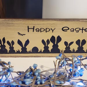 Può includere: Un cartello di legno con una finitura invecchiata di colore marrone chiaro. Il cartello recita "Happy Easter" in lettere nere.  Sagoma di coniglietti e uova sono dipinte sul cartello.
