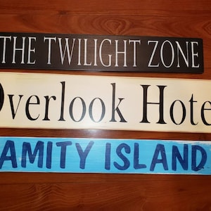 Op de afbeelding: Drie houten borden met geschilderde tekst. Het bovenste bord is zwart met witte tekst die "THE TWILIGHT ZONE" luidt. Het middelste bord is cr&egrave;mekleurig met zwarte tekst die "Overlook Hotel" luidt. Het onderste bord is blauw met donkerblauwe tekst die "AMITY ISLAND" luidt.