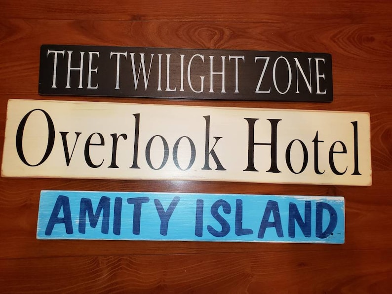 Op de afbeelding: Drie houten borden met geschilderde tekst. Het bovenste bord is zwart met witte tekst die "THE TWILIGHT ZONE" luidt. Het middelste bord is cr&egrave;mekleurig met zwarte tekst die "Overlook Hotel" luidt. Het onderste bord is blauw met donkerblauwe tekst die "AMITY ISLAND" luidt.