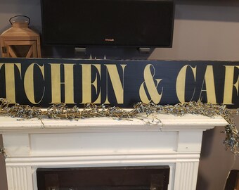 Könnte beinhalten: Ein rechteckiges schwarzes Holzschild mit den Worten "KITCHEN & CAFE" in goldfarbenen Buchstaben. Das Schild wird über einem weißen Kaminsims mit einer dekorativen Girlande und einer Holzlaterne angezeigt.