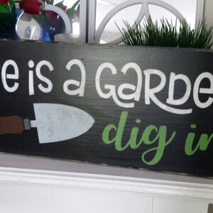 Op de afbeelding: Zwart houten bord met witte tekst "Life is a GARDEN dig in". Een bruine en zilveren troffelafbeelding staat eronder. Het bord is een decoratief item voor een tuin of huis.