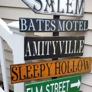 Op de afbeelding: Een houten bord met meerdere richtingspijlen die naar verschillende locaties uit horrorfilms wijzen. De borden zijn in verschillende kleuren geschilderd en bevatten de namen: Salem, Bates Motel, Amityville, Sleepy Hollow, Elm Street en  'NSYLVANiA.