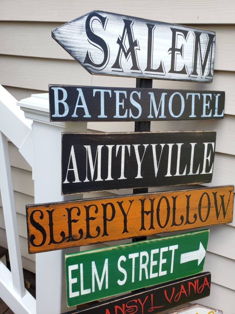 Op de afbeelding: Een houten bord met meerdere richtingspijlen die naar verschillende locaties uit horrorfilms wijzen. De borden zijn in verschillende kleuren geschilderd en bevatten de namen: Salem, Bates Motel, Amityville, Sleepy Hollow, Elm Street en  'NSYLVANiA.