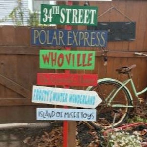 Op de afbeelding: Een houten wegwijzer met meerdere borden die in verschillende richtingen wijzen. De borden zijn in verschillende kleuren geschilderd en tonen kerstbestemmingen, waaronder "Noordpool", "Kerststad", "34e Straat", "Pool Express", "Whoville", "Frosty's Winter Wonderland" en "Eiland van de misfit speelgoed".