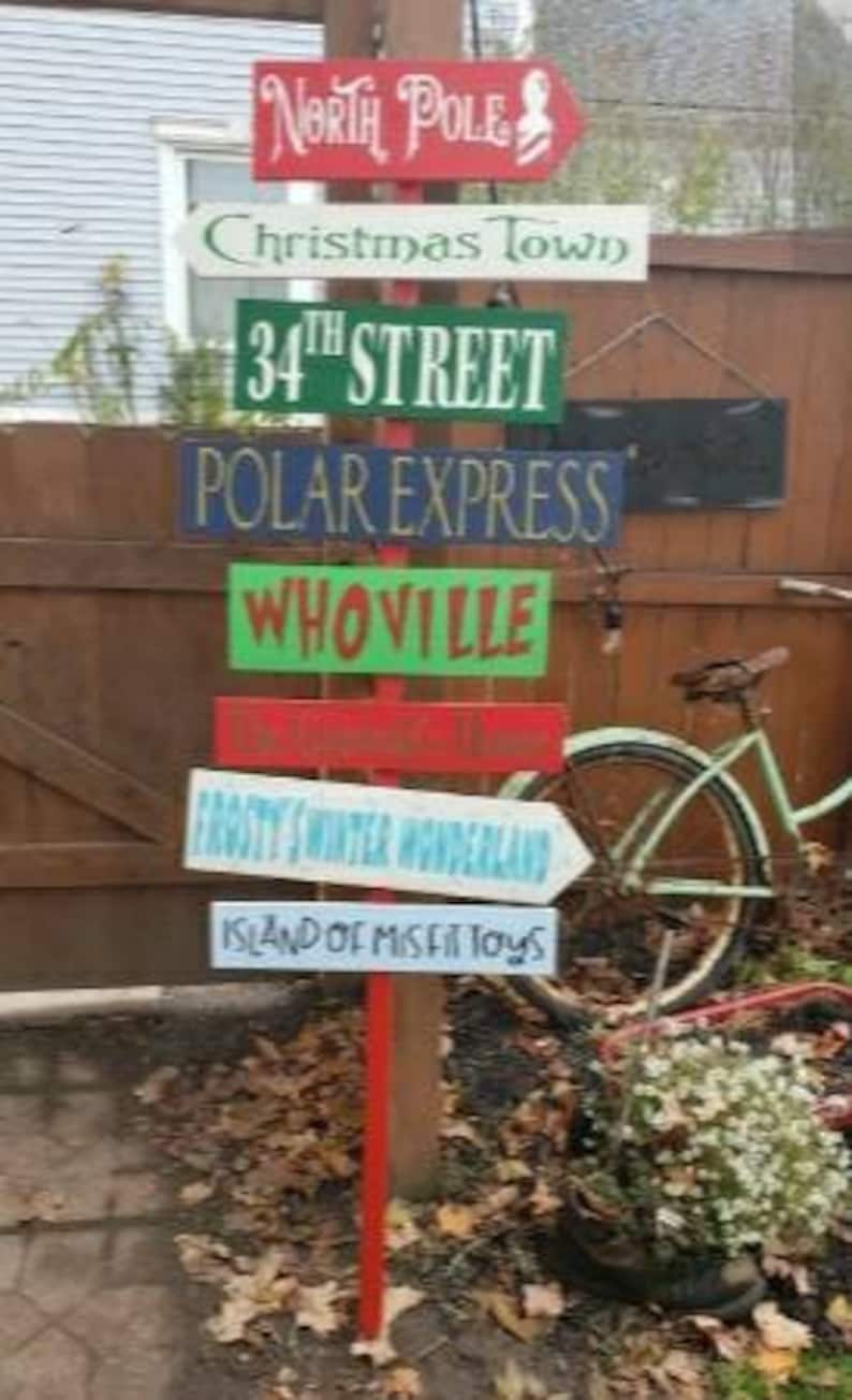 Op de afbeelding: Een houten wegwijzer met meerdere borden die in verschillende richtingen wijzen. De borden zijn in verschillende kleuren geschilderd en tonen kerstbestemmingen, waaronder "Noordpool", "Kerststad", "34e Straat", "Pool Express", "Whoville", "Frosty's Winter Wonderland" en "Eiland van de misfit speelgoed".