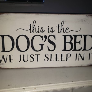 Puede incluir: Un letrero rectangular de madera con fondo blanco desgastado y letras negras. El texto dice "this is the DOG'S BED WE JUST SLEEP IN IT". El letrero tiene bordes marrones y es probablemente para la decoración del hogar.
