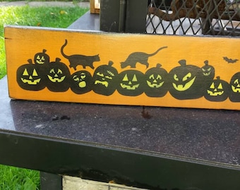 May include: A wooden sign with a Halloween theme. It features a row of black silhouette jack-o'-lanterns with yellow faces and two black cats. The sign is painted orange.