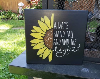 May include: Black wooden sign with a yellow sunflower painted on the left side and the text "ALWAYS STAND TALL AND FIND THE light" in white on the right side.