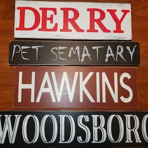 Op de afbeelding: Vier houten borden met verschillende namen van steden erop geschilderd. Het bovenste bord zegt "DERRY" in rode letters op een witte achtergrond. Het tweede bord zegt "PET SEMATARY" in witte letters op een zwarte achtergrond. Het derde bord zegt "HAWKINS" in witte letters op een rode achtergrond. Het onderste bord zegt "WOODSBORO" in witte letters op een zwarte achtergrond.