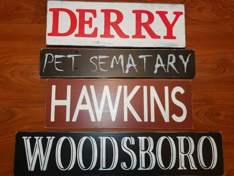Op de afbeelding: Vier houten borden met verschillende namen van steden erop geschilderd. Het bovenste bord zegt "DERRY" in rode letters op een witte achtergrond. Het tweede bord zegt "PET SEMATARY" in witte letters op een zwarte achtergrond. Het derde bord zegt "HAWKINS" in witte letters op een rode achtergrond. Het onderste bord zegt "WOODSBORO" in witte letters op een zwarte achtergrond.